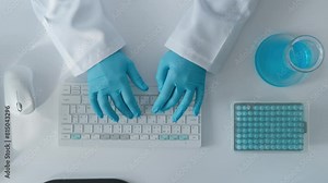 Hands in medical gloves and a doctor's gown typing on a computer keyboard. White table of a modern laboratory close-up view from above.