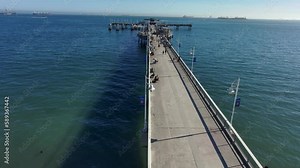 aerial footage of a long concrete pier over the vast deep blue ocean water with people walking along the pier and curved light posts along the pier at Belmont Veterans Memorial Pier in Long Beach
