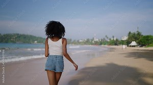 Multiethnic black woman runs on ocean beach on tropical vacation at exotic island. African american female turns, flirts running on sea shore with straw hat. Bipoc has fun on travel tour. Follow me.