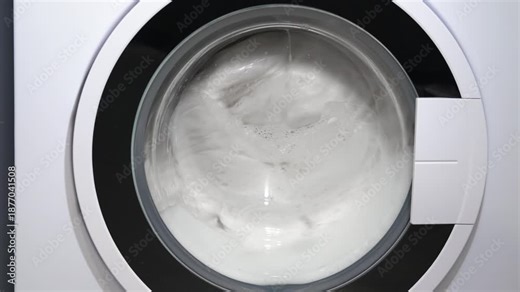 Washing machine spinning white laundry close-up. Front-loading washing machine spinning clean white clothes during the rinse and spin cycle, with soapy water being gradually drained away