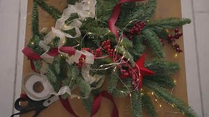 top view plan. Fully visible the table with the decorations. Female hands put and finalize Christmas gift wrapped in craftool paper on a wooden table. Bandaging tape and tied bow.