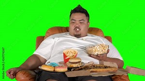 Greedy obese man eating popcorn, pizza, donuts, and burger on the sofa. Shot in 4k resolution with green screen background