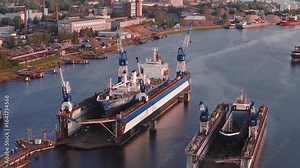 A shipyard in Riga, Latvia features cargo ships in dry docks, cranes in motion, industrial buildings, and warm sunlight reflecting on calm water.