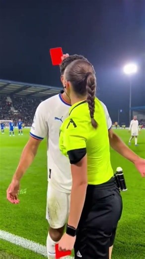female referee halts the match and firmly raises a red card toward a male player