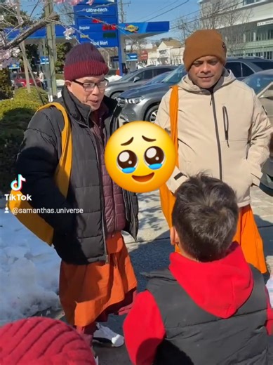 Buddhist monks standing on a sidewalk near a gas station and a modern white municipal building in Takoma Park, just across the border from Washington, D.C.. Visible clues include a Sunoco station sign, Maryland license plates on nearby cars, and a white civic building consistent with the Takoma Park Community Center. The cherry blossoms in bloom suggest the footage was taken during spring in the D.C. region. The men in orange robes are Buddhist monks. Their clothing style is typical of monks fro