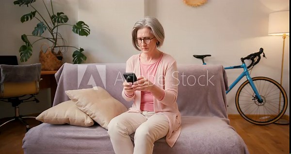 Mature old 60s woman, older middle aged female customer holding smartphone using mobile app, texting message, search ecommerce offers on cell phone technology device sitting on couch at home