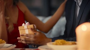 A diverse couple sits closely together at a table in a cozy restaurant, sharing a romantic dinner. The woman hands a beautifully wrapped gift to her partner, filled with affection.