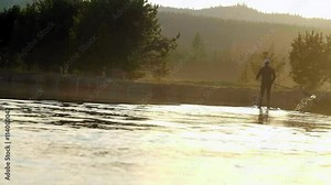 From a wide shot with a slightly canted angle, a man in a wetsuit paddleboards down an Oregon river away from camera during a beautifully glorius sunset