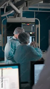 An experienced doctor oversees two fellow surgeons in the operating room. Monitors with patient disease data and X-rays in the observation room. Vertical shot.