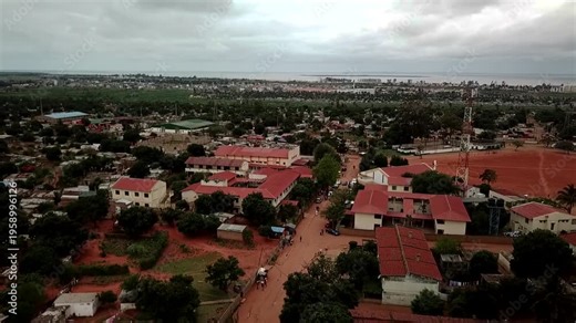 Aerial view of a suburban neighborhood in Maputo, Mozambique, with red‑roofed houses, dirt roads, scattered trees, and the distant city skyline and bay under an overcast sky.
