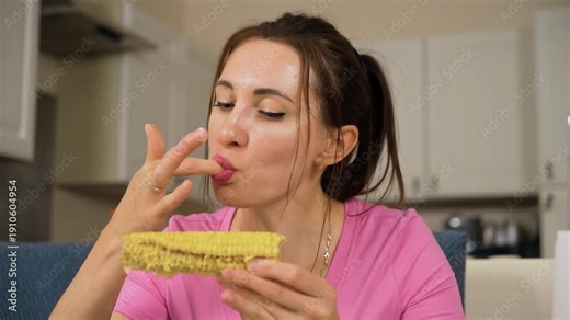 Female licking finger after eating corn cob at table. Woman enjoying taste of maize while holding cob in hand. Girl savoring sweetcorn and cleaning hand during meal in kitchen