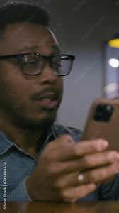 Vertical shot of African American man rejoices at the soccer team victory in championship, checks bookmaker ratings online using phone. Happy sport fan wins bet on football match sitting in bar or