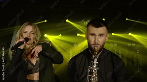 Musical band group of two people playing song, performing on concert musician stage with lights. Shooting a music video. Singer vocalist girl, saxophonist sax man. Actors dancing, singing. Slow motion