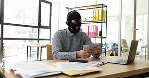 Internet operator scammer in a balaclava with headphones with microphone counts money at computer desk. Cybercrimes and financial crimes online