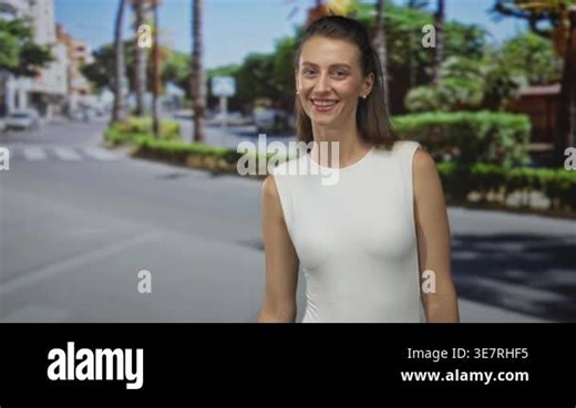 Woman making ok sign with both hands while smiling and winking, wearing sleeveless white top on sunlit street with palm trees; optimism approval Stock Video Footage - Alamy