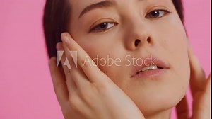 Young beautiful woman with clean skin close up touching her cheekbones and cheeks with both hands while looking at the camera on a pink background. Concept of skin care for health and beauty