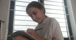 Smiling smart little girl writes in a notepad. Cute preteen school girl pupil studying at home, teenage girl expressing lonely thoughts while sitting on bed in her room. 4K