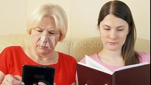 Mother reading e-book and daughter reading physical print book. Concept of paper vs digital reading, e-books replacing paper books. Concept of sharing reading with your kids