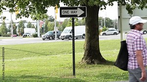 one way white and black horizontal rectangle arrow sign on metal post under tree with road street behind with cars vehicles traffic passing and pedestrian passing in front