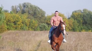 Athletic man with a pumped body rides a horse across the field towards the camera