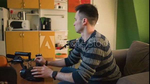 Person sitting on sofa in home studio, focusing on video camera setup with tripod, preparing for filming project. Workspace with coffee machine and colorful objects in background