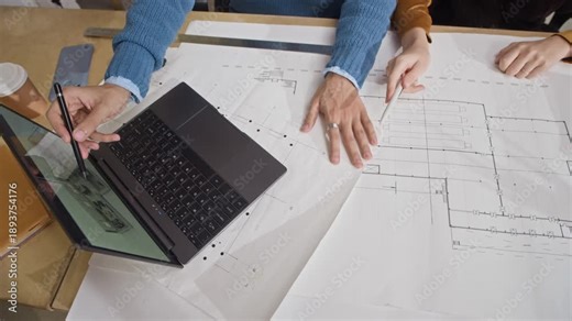 Top down view of two unrecognizable colleagues working together on laptop displaying rotating digital 3d house exterior model while reviewing floor plan at table in contemporary architectural office