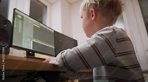 A young boy sits at a desk using a computer with dual monitors, immersed in learning and coding. He is wearing a striped shirt and appears focused on the screen.