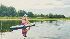 Woman sitting on sup board in purple swimsuit floating on lake, river rowing with oar, back view. Outdoors activity on nature. Female paddling on SUP board. Active sport vacations, adventure concept.