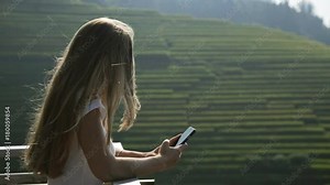 Young attractive woman watching sunrise over Longji Rice Terraces from top of the mountain. Blonde beautiful girl takes panorama photos on phone.