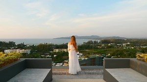 Back View of Alone Woman Standing on Rooftop on Sunset with Seaview. Pensive Woman Touch Her Long Hair in White Dress on Terrace Looking into Distance and Thinking about Future. Travel Concept