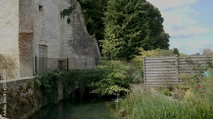 Pan-Slow moving River Coln flowing by stone cottage Bibury, a village and civil parish in Gloucestershire, England