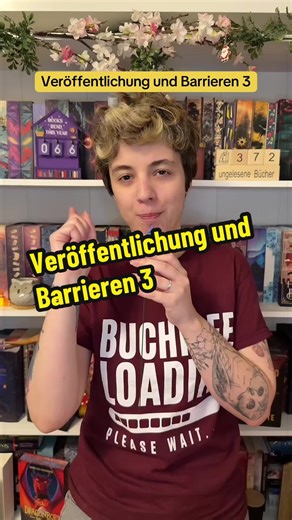 Part 1: @books_and_awareness Part 2: @books_and_awareness Barrieren bei der Veröffentlichung mit Blindheit Teil 3 Nadine mit kurzem, hellem Haar und tätowierten trägt ein weinrotes T-Shirt mit der Aufschrift „Buchidee loading... please wait“. Nadine hält ein kleines, blaues, schmetterlingsförmiges Mikrofon in der Hand und steht vor einem Regal, das mit einer Vielzahl von Büchern gefüllt ist. Über dem Regal sind weiße Blumen als Dekoration angebracht. #BookTokGermany #AutorenAufTikTok #Autorinnen