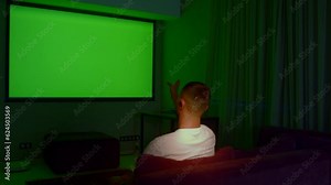 Man watching TV with green screen in living room, back view. Media. Man switching channels with hand gestures, gesture interface, modern technology.