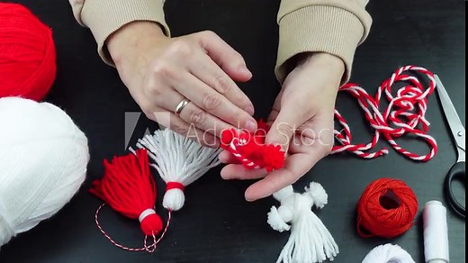 Woman making handmade traditional martisor, from red and white strings with tassel. Symbol of holiday 1 March, Martenitsa, Baba Marta, beginning of spring in Romania, Bulgaria, Moldova