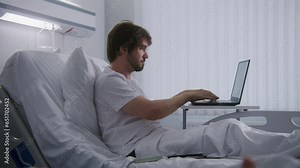 Adult man lies on bed in hospital ward, remotely works using laptop. Programmer works in medical center and chats using phone. Patient fully recovering after successful surgery. Modern bright clinic.