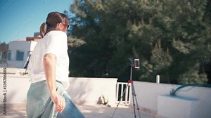 A young attractive woman doing online dance class for the social network using a smartphone on a tripod on the roof with sea view. Streaming during the self-isolation time.