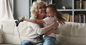 Happy pleasant elderly senior grandmother in glasses holding joysticks, playing online video games on tv with overjoyed little preschool child granddaughter, celebrating winning competition at home.