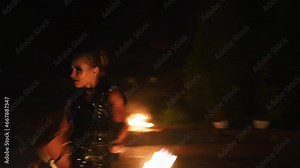 Fire girl spins burning poles at the night fire performance. Female artist juggling torches with flame during fire show at venue for tourists. Young woman working with fire on the street festival.