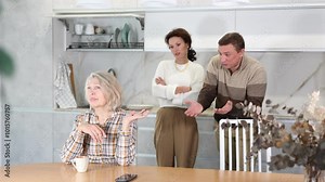 Upset old woman sitting at table with her back to angry middle-aged man and woman quarreling to her in the kitchen. High quality 4k footage