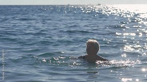Elderly senior woman with gray hair, swimming in sea near shore, afternoon sun reflects on water surface behind