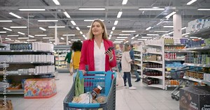 Beautiful Female Shopper Pushes a Cart Through the Supermarket, Holding Her Smartphone in One Hand. She Is Checking Her Grocery List and Comparing Prices Online as She Navigates the Aisles