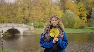 Woman with curly hair and a cozy scarf holding yellow autumn leaves by a scenic lake, with a stone bridge and vibrant fall foliage in the background. Romantic walk through the autumn park.