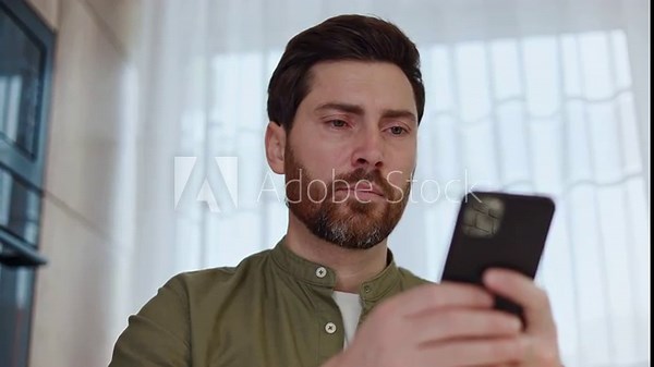 Serious male freelancer with trendy hairstyle using application in modern smartphone for browsing internet. Caucasian bearded man searching online new distance work while sitting at domestic office.