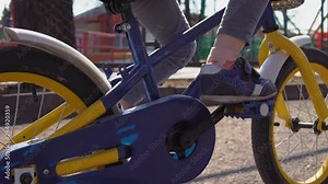 Camera follows little girl pedaling blue bicycle with yellow basket. Child is driving along an old asphalt path in park on sunny day.