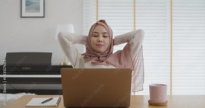 Young islam girl workforce sit easy watch seminar at cozy home. Asia woman people work life balance relax smile lean bending back hand behind head look at laptop remote learn online MBA college class.