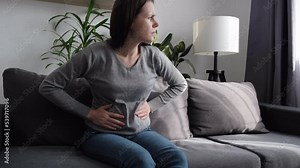 Upset female sitting on couch suffering from indigestion flatulence abdominal problem concept. Sick young woman in pain holding belly stomach feeling hurt abdomen ache gastritis pancreatitis symptoms