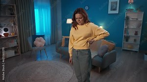 A woman suffers from back pain while standing in the living room. A woman massages her lower back, arches her back. A woman suffers from pain in lower back, pinched nerve. Home medicine concept.