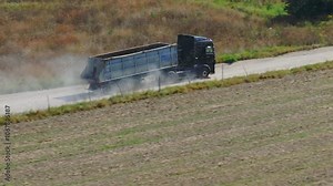 Large lorry is driving on a dusty country road, leaving a trail of dust behind. The lorry is surrounded by fields and trees