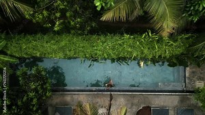 Drone top view woman in bikini lounging in crystal-clear pool surrounded by greenery. Sips cocktail, enjoying solitude in tropical paradise. Sunlight filters through palms, reflecting on water.