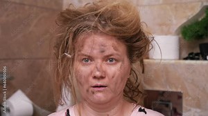 Blonde woman with unkempt hair sits in tiled toilet after small explosion. Female looks at one spot in bathroom with surprised and shocked expression